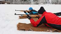 Zwei Sportler liegen mit Gewehr im Anschlag auf einer Matte am Boden im Schnee