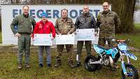 Fünf Menschen schauen in die Kamera, drei halten Schecks. Im Vordergrund ein Kindercrossmotorrad.