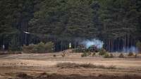 Ein Gefechtsfahrzeug in der Form eines Panzers bei der Schussabgabe im Gelände. Rauch steigt auf.