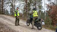 Ein Motorradfahrer mit Warnweste steht vor einem Ausbilder auf einem ansteigenden Waldweg