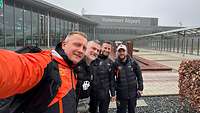 Vier Persaonen in einem Invictus Games Trainingsanzug stehen vor einem Terminal an einem Flughafen.