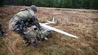 Ein Soldat bückt sich auf einer Wiese und hebt eine weiße Drohne in der Form eines Flugzeugs.