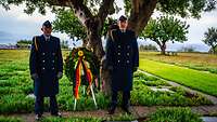 Zwei Soldaten in blauer Uniform stehen mit einem Kranz unter einem Olivenbaum.