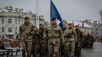 Ein Soldat marschiert an der Spitze einer Gruppe Soldaten. Hinter ihm weht die NATO Flagge. 