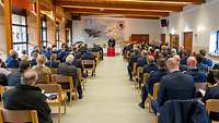 Ein langer Raum mit einem Altar in der Mitte; viele Menschen sitzen und hören einem Pfarrer zu.