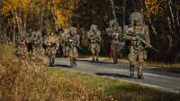 Eine Gruppe von Soldaten in Flecktarnuniformen mit Gepäck und Waffen geht eine Straße entlang.