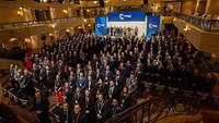 Viele Soldaten und Zivilisten stehen vor einer Bühne zwischen Sitzplätzen in einem Konferenzsaal