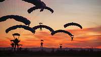 Soldaten gleiten mit Fallschirmen vor dem Hintergrund eines glutroten Sonnenaufgangs gen Erdboden.