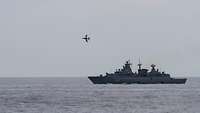 Ein Flugzeug überfliegt ein graues Kriegsschiff auf offener See.