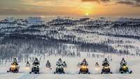 Soldaten im Schneetarn fahren auf Motorschlitten nebeneinander durch Schnee. Die Sonne geht unter.