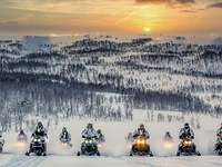 Soldaten im Schneetarn fahren auf Motorschlitten nebeneinander durch Schnee. Die Sonne geht unter.