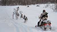 Soldaten im Schneetarnanzug fahren auf Motorschlitten auf einem verschneiten Feld in Richtung Wald.