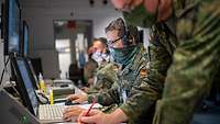 Vier Soldaten sitzen mit Mund-Nasen-Schutz in einem Raum und arbeiten am Computer.