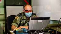 Soldat mit Mund- Nasen-Schutz und Schutzbrille arbeitet an Computer.