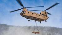 Eine CH-47 in der Luft. Unter ihr ist eine Außenlast mit Seilen an einem Haken befestigt.