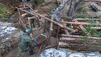 Soldaten bauen ein Gestell aus Rundhölzern in einem Graben. Ein Soldat schiebt eine Schubkarre mit Erde.