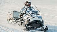 Zwei Soldaten fahren auf einem Motorschlitten mit Anhänger durch eine verschneite Landschaft.