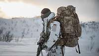 Ein Soldat im Schneetarnanzug steht mit seinem riesigen Rucksack auf einer verschneiten Fläche.