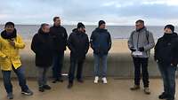 Am windigen Nordseestrand hält ein Mann einen Vortrag vor einer Gruppe anderer Männer.