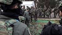 Soldaten stehen um ein Miniaturmodell des Geländes. Im Hintergrund befinden sich ein weißes Zelt und Wald.