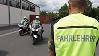 Soldat in Warnweste mit Aufschrift "Fahrlehrer", im Hintergrund zwei Fahrschüler auf Motorrädern