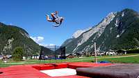 Ein junger Mann springt auf einem Trampolin vor einem Panorama aus Bergen durch die Luft.