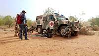Malische Soldaten versorgen einen verwundeten Soldaten vor einem gepanzerten Fahrzeug, andere Soldaten sichern die Umgebung