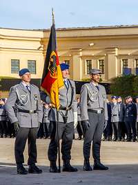 Three service men in basic dress, the one in the middle holds the flagpole with the Federal Institutions Flag