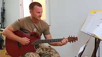 Ein Soldat sitzt mit einer Gitarre in einem Raum und musiziert.