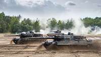 Three battle tanks in staggered formation speed across a sandy training area. Dust is whirled up.