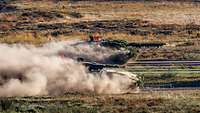 A Leopard 2 battle tank and a Puma infantry fighting vehicle move rapidly side by side across a training area. Dust swirls. 