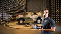 A man in civilian clothing with a measuring device stands next to a combat vehicle in a soundproofed workshop. 