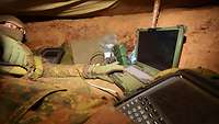 In a concealed position underground, a soldier with IT equipment operates a laptop computer.