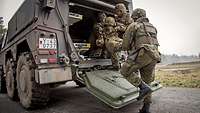 Three soldiers carry an injured comrade into a military vehicle whose rear door is open.