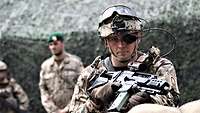 A soldier with the digital “Future Infantryman” system and a G36k rifle stands in front of a camouflage net.