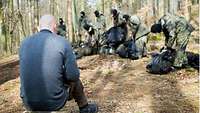 Soldiers put down their backpacks in the forest. In the foreground, a soldier sits on a tree stump.