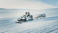 Two soldiers ride a snowmobile towing a cargo sled down a snowy slope.