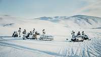 Several soldiers mounted on snowmobiles towing cargo sleds move through a landscape completely blanketed in snow.