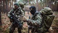 Two soldiers with balaclavas, packs and weapons stand in a forest area: One briefs the other.