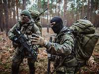 Two soldiers with balaclavas, packs and weapons stand in a forest area: One briefs the other.