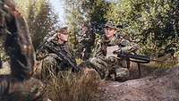 Two servicewomen crouch in wooded terrain with their weapons, conferring with each other.