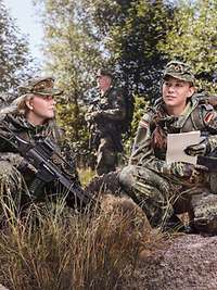 Two servicewomen crouch in wooded terrain with their weapons, conferring with each other.