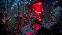 Several soldiers are assembled around an armoured vehicle. The rear door is open; red light illuminates the vehicle’s interior.