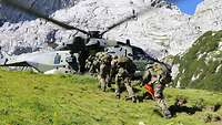 Mehrere Soldaten laufen in Reihe zu einem Hubschrauber auf einer Wiese vor einer Felswand.