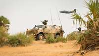 Ein Dingo steht in der malischen Wüste und eine Transall fliegt darüber hinweg