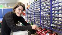 Eine Frau mit Brille sitzt an einem Arbeitsplatz in einer Elektriker-Werkstatt, an der Wand viele kleine Schubladen.