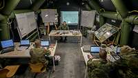 Vier Soldaten im logistischen Gefechtsstand an den Computern. Im Hintergrund sind Karten und Whiteboards zu sehen.