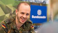 Ein Soldat im Gespräch mit einer Person, im Hintergrund ist das das Logo der Bundeswehr zu sehen.