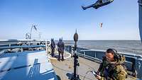 Ein Mann in Uniform steht auf einem Schiff neben einem Mikrofon, im Hintergrund ein Hubschrauber.