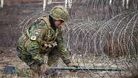 Ein Soldat hockt an einem Zaun aus Stacheldraht und schiebt eine Metallstange zwischen die Drähte.